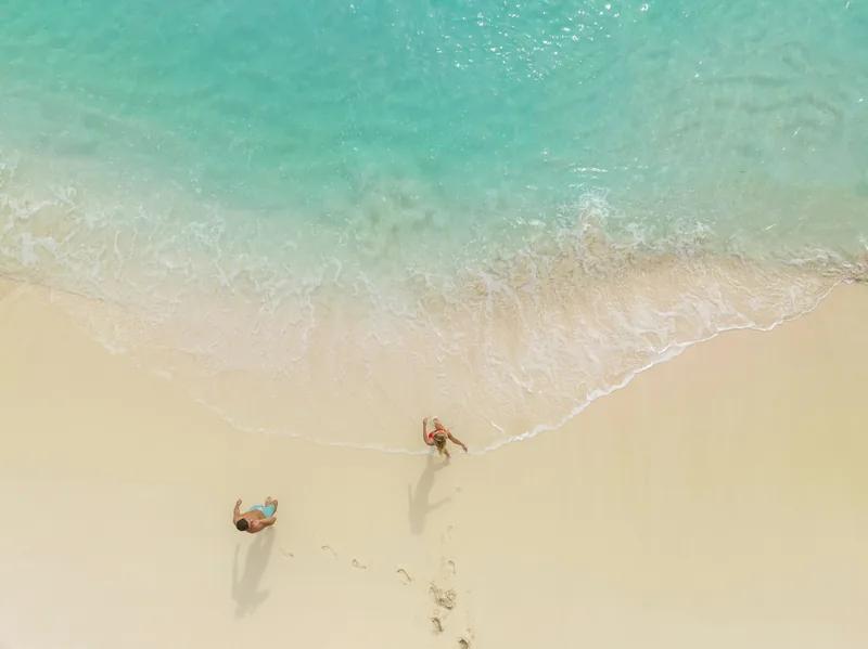 Two figures walking at the water's edge on a pale beach, seen from directly above