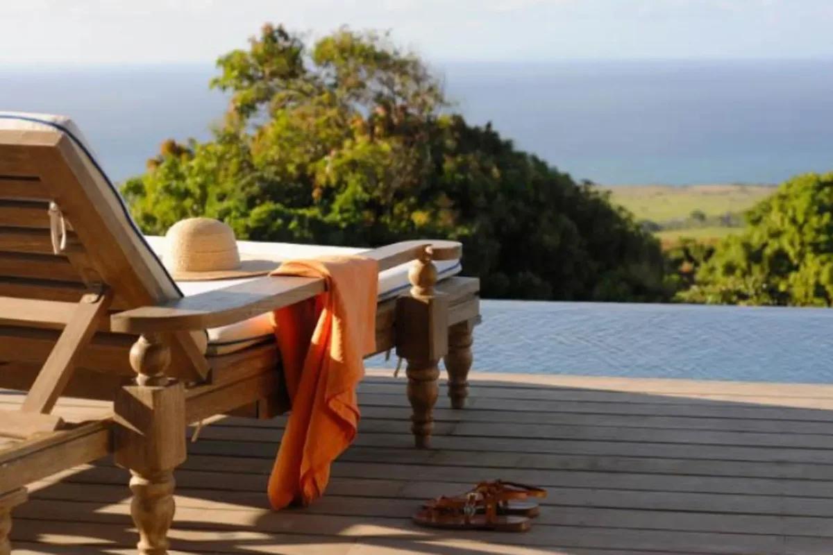 A straw hat and orange towel on a timber lounger on a deck above green hillside and sea